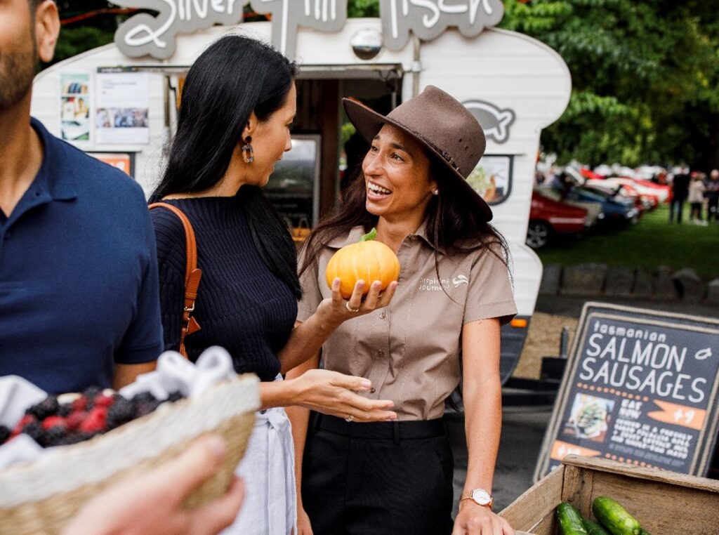 Two women with long dark hair, one holding a mango and the other with a brown hat and uniform of a travel director at a market stall